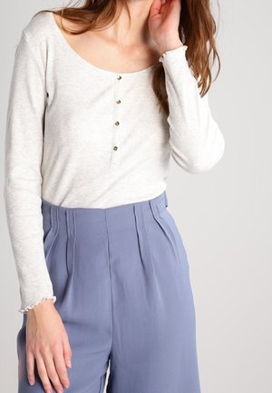 Femme portant un haut en maille côtelée crème à manches longues avec boutons, associé à un pantalon bleu clair plissé à taille haute, sur un fond uni.