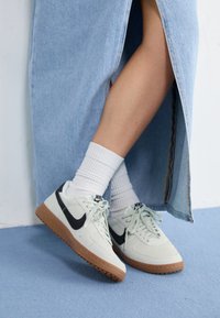 Light grey suede sneakers with black accents and white laces, featuring a brown rubber sole. Worn with white ribbed socks and a denim skirt.