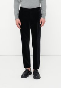 Osoba oblečená v čiernych nohaviciach s rovnými nohavicami, čiernych slip-on topánkach a šedom tričku s dlhým rukávom, stojí na bielom pozadí.