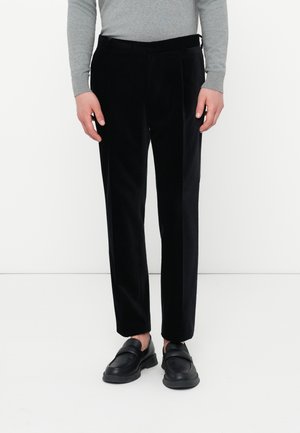 Osoba oblečená v čiernych nohaviciach s rovnými nohavicami, čiernych slip-on topánkach a šedom tričku s dlhým rukávom, stojí na bielom pozadí.