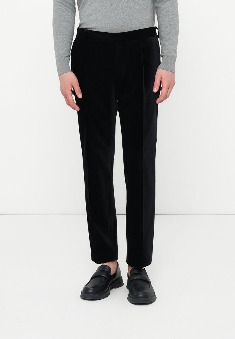 Osoba oblečená v čiernych nohaviciach s rovnými nohavicami, čiernych slip-on topánkach a šedom tričku s dlhým rukávom, stojí na bielom pozadí.