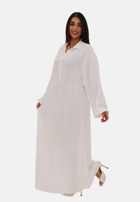 Robe longue blanche en tissu texturé, dotée d'un col à boutons, de manches longues et d'une coupe ample. Accentuée par des sandales à talons.