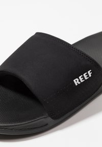 Sandale noire à semelle en caoutchouc texturé, avec une sangle douce et rembourrée comportant des perforations et le logo "REEF" en blanc.