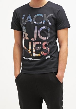 Personne portant un T-shirt noir à manches courtes avec un grand texte multicolore "JACK & JONES ORIGINALS 1975", mains dans les poches d'un pantalon noir.