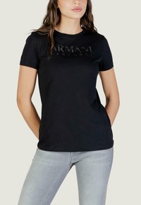 Vrouw die een zwarte Armani Exchange T-shirt met korte mouwen en lichtgrijze spijkerbroek draagt, staand met de handen achter haar rug.