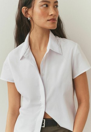 Biała koszula na krótki rękaw z guziczkami, o wyostrzonym kołnierzu i lekko luźnym kroju, zestawiona z ciemnym paskiem. Gładka faktura, bez wzorów.