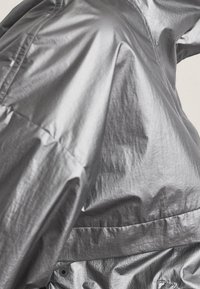 Glänzender, metallischer Stoffmantel mit sichtbaren Nähten und Falten, der die Textur und die reflektierende Oberfläche in Nahaufnahme hervorhebt.