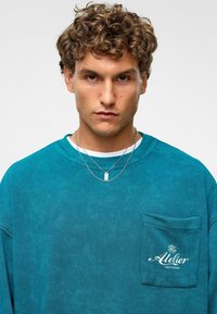 Teal langemouw shirt met een gestructureerde afwerking, voorzien van een borstzak versierd met een wit "Atelier" logo en gelaagde zilveren kettingen.