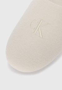 Beige papucs sima filc textúrával, a felületen kiemelkedő, hímzett "cK" logóval, azonos színű fonallal.