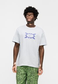 Ανοιχτό γκρι βαμβακερό μπλουζάκι με κεντρικό μοβ λογότυπο Nike, κοντά μανίκια και λεπτομέρειες με σχέδια στο πλάι. Συνδυάζεται με πράσινο παντελόνι με σχέδιο πιτσιλιές.