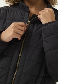 Chaqueta acolchada negra con un collar alto, que presenta un zipper dorado y una textura acolchada. Manos ajustando el zipper.