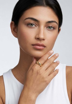 Sötét hajú, zöld szemű nő felemeli az állát, fehér ujjatlan felsőt visel és vastag aranygyűrű van az ujján.