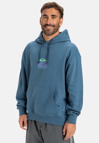 Hoodie en coton bleu avec une poche frontale, capuche ajustable à cordon, et un graphique de logo coloré sur la poitrine. Texture douce, coupe décontractée.