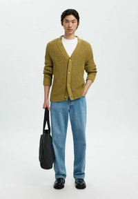 Ung man som står, iklädd en olivgrön stickad kofta, vit t-shirt, blå jeans, svarta skor, håller en svart tote-väska i vänster hand.