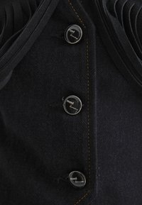 Schwarzer Denim-Stoff mit auffälligen Knöpfen und metallischen Akzenten. Das Oberteil hat ein geschichtetes Design mit strukturierten Details und kontrastierenden gelben Nähten.