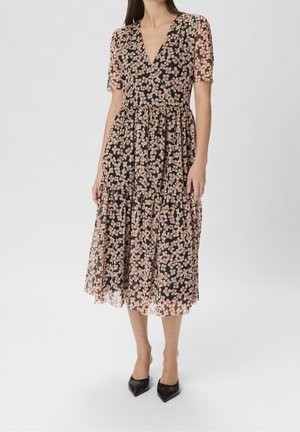 Frau trägt ein schwarzes Midikleid mit rosa Blumenmuster, V-Ausschnitt, kurzen Ärmeln und plisziertem Rock, kombiniert mit schwarzen spitzen Schuhen.