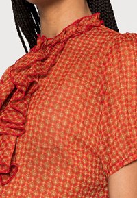 Blusa rossa a fantasia con colletto e rifiniture arricciate. Il tessuto è trasparente e texturizzato, con un motivo a diamante in tonalità più chiare.