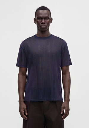 Uomo dalla pelle scura che indossa una maglia lavorata a maglia blu navy a maniche corte con motivo e pantaloni scuri, in piedi davanti a uno sfondo bianco semplice.