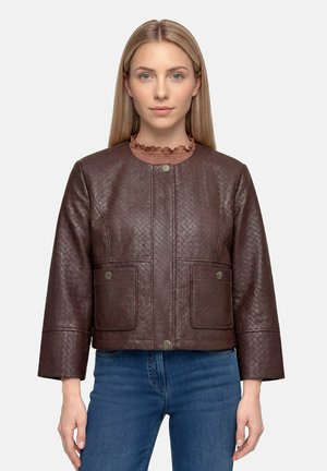 Femme aux cheveux blonds et raides porte une veste marron à motifs avec des poches à pressions et un jean bleu sur un fond uni.