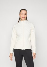 Weiße Zip-Jacke mit gerippten Ärmeln und gesteppter Vorderseite. Merkmale einen hohen Kragen und eine figurnahe Passform, kombiniert mit schwarzen Hosen.