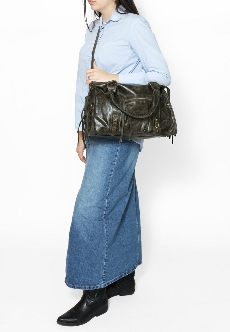 Borsa in pelle di colore verde scuro, design rettangolare con dettagli intrecciati, fibbie dorate e due opzioni di trasporto. Modella che indossa una camicia azzurro chiaro e una gonna in denim.