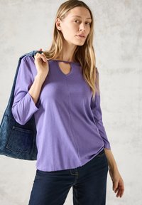 Maglietta a maniche lunghe viola con scollatura a chiave, leggera e con vestibilità rilassata, abbinata a jeans di denim scuri e una giacca di denim.