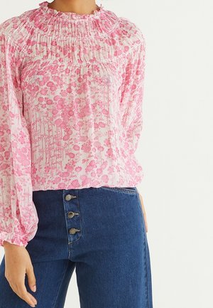 Vrouw draagt een roze bloemenlange mouwen blouse met gerimpelde halslijn en een hoge taille blauwe jeans met drie zichtbare knopen.