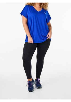 Kvinde iført en løs, blå V-hals T-shirt, sorte leggings og marineblå sportssko, stående mod en hvid baggrund.