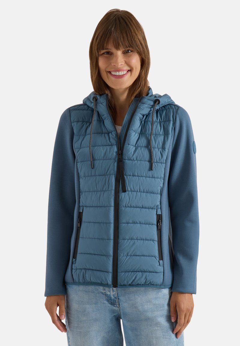 Femme souriante portant une veste matelassée bleue à capuche avec cordons et un jean bleu clair sur un fond uni.