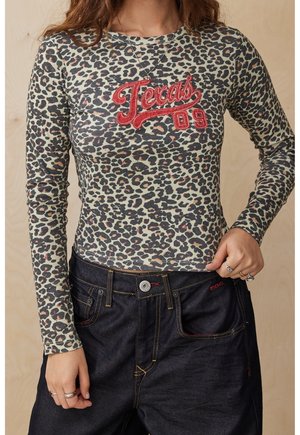 Femme portant un haut à manches longues imprimé léopard avec un texte rouge "Texas 09" et un jean denim bleu foncé avec des coutures jaunes.