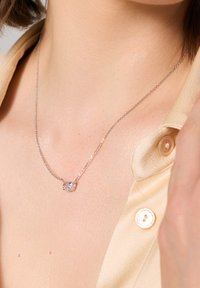 Srebrny naszyjnik łańcuszkowy z wiszącym owalnym przezroczystym kamieniem szlachetnym, noszony na odkrytej skórze, z widoczną satynową beżową kołnierzykiem bluzki.