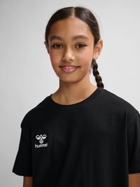 Junges Mädchen mit geflochtenem Haar, lächelnd, trägt ein schwarzes T-Shirt mit weißem hummel-Logo auf einem einfarbigen hellgrauen Hintergrund.