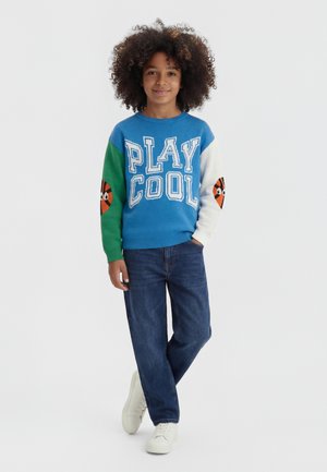 Bambino con capelli ricci che indossa un maglione blu con la scritta "PLAY COOL", maniche diverse con volti di cartoni animati, jeans blu e scarpe da ginnastica bianche.