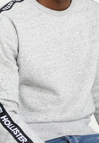 En person som bär en ljusgrå sweatshirt med texten "HOLLISTER" i svart och vitt på vänster ärm, sitter med blå jeans synliga.