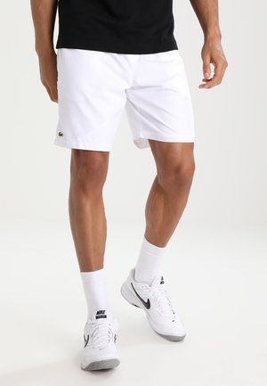 Man die witte Lacoste-short draagt, een zwart shirt, witte Nike-sokken en witte Nike-sneakers, staand tegen een witte achtergrond.