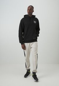 Felpa nera con cappuccio, tasca frontale e logo; abbinata a pantaloni della tuta grigi chiari con strisce laterali nere; sneakers nere e gialle.
