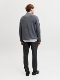 Άνδρας με την πλάτη, φοράει γκρι πουλόβερ πάνω από ριγέ πουκάμισο, σκούρο παντελόνι και μαύρα επίσημα παπούτσια σε απλό λευκό φόντο.