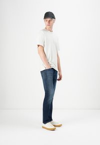Young man wearing a black cap, white t-shirt, blue jeans, and white sneakers standing with one hand in pocket against white background.