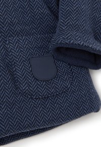 Tessuto a spina di pesce blu navy con una morbida fodera in pile e un dettaglio a toppa testurizzata. Il design presenta sottili motivi diagonali.