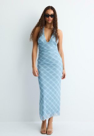 Donna con lunghi capelli ricci che indossa occhiali da sole neri, un abito smanicato a quadri blu e tacchi con cinturini trasparenti, in piedi davanti a uno sfondo bianco.