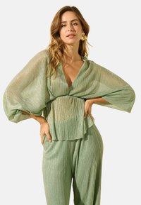 Haut plissé léger vert clair, transparent, avec des manches larges et un décolleté en V profond, associé à un pantalon plissé assorti. Texture brillante notable sur l'ensemble.