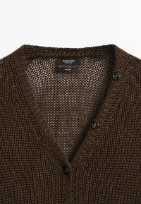 Cardigan en lin marron avec un tricot texturé, présentant un col en V et deux boutons noirs. Les étiquettes indiquent la marque et la composition du matériau.