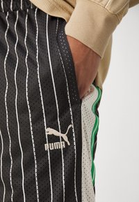 Shorts de sport Puma en maille noire et grise avec des rayures blanches verticales, dotés d'une poche latérale et d'un accent vert le long de la couture.