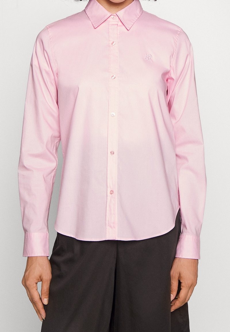 Femme portant une chemise rose clair à manches longues boutonnée avec col et un pantalon noir, debout devant un fond clair uni.