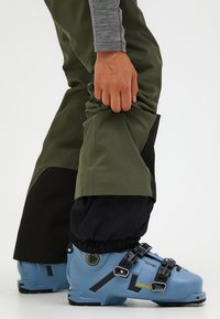 Skischoenen in lichtblauw met zwarte hardware en banden; groene en zwarte broek met een gladde textuur en aansluitende boorden over de schoenen.