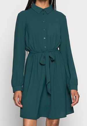 Woman wearing a teal long-sleeve button-up dress with a collar and a tied waist belt, standing against a plain background.