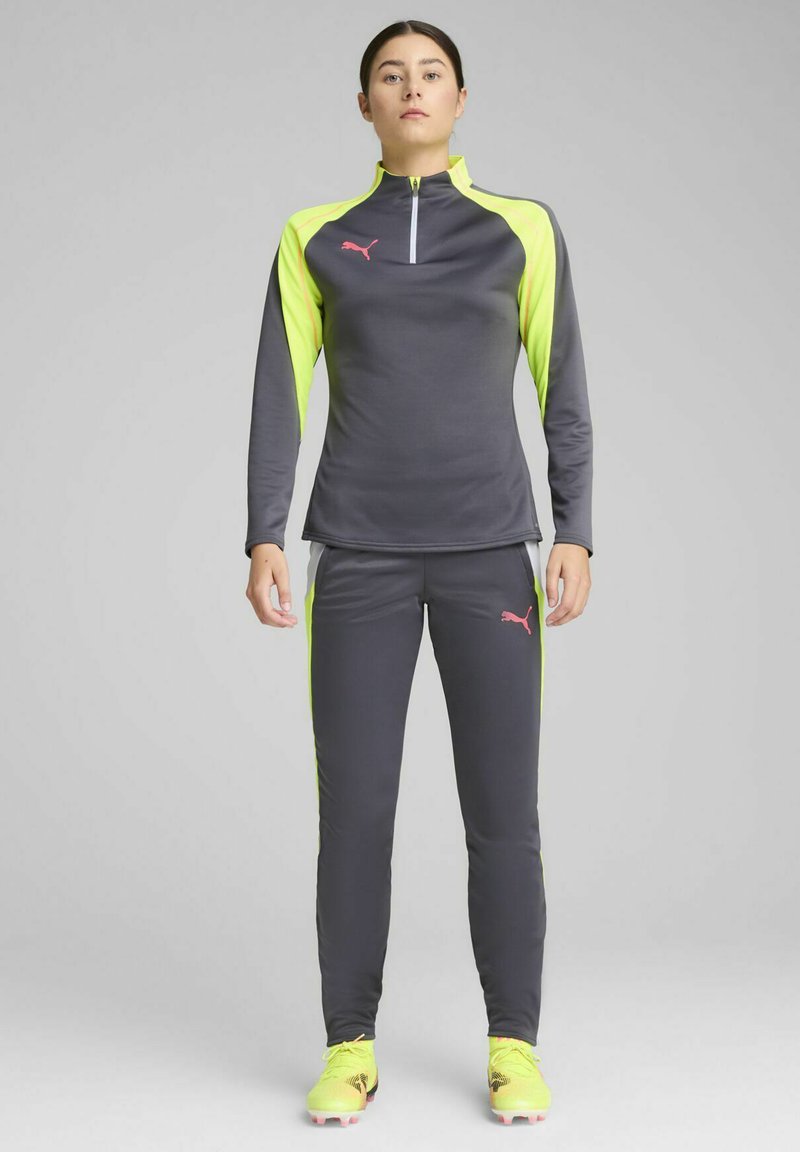 Sivi sportski top s dugim rukavima i visokim ovratnikom, naglašen svijetlosmeširim stranicama, uparen s odgovarajućim sivim hlačama. Detalj s ružičastim logotipom.