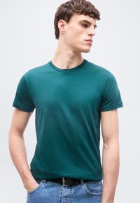 Νεαρός άνδρας που φοράει απλό σκούρο πράσινο-μπλε κοντομάνικο t-shirt και ανοιχτό γαλάζιο τζιν, κοιτάζει προς τα δεξιά του με το ένα χέρι στην τσέπη.