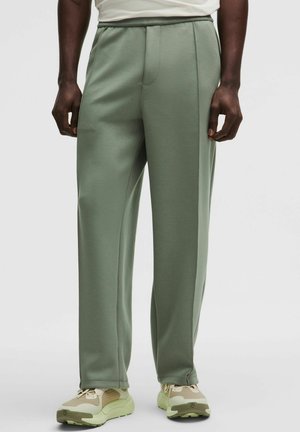 Egy férfi világoszöld, egyenes szárú nadrágot és bézs sportcipőt visel, egyszínű fehér háttér előtt áll.