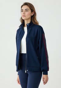 Giacca zip in morbido tessuto di colore navy, con colletto alto e dettagli a righe rosse sulle maniche. Texture liscia e vestibilità comoda.